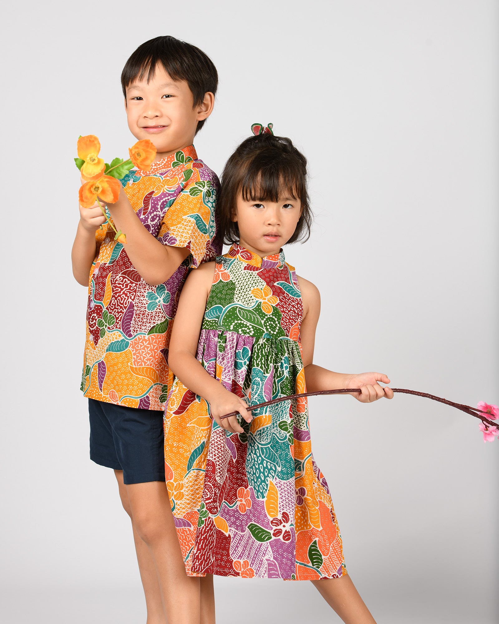 Two children in matching colorful batik outfits holding toy flowers again a plain background