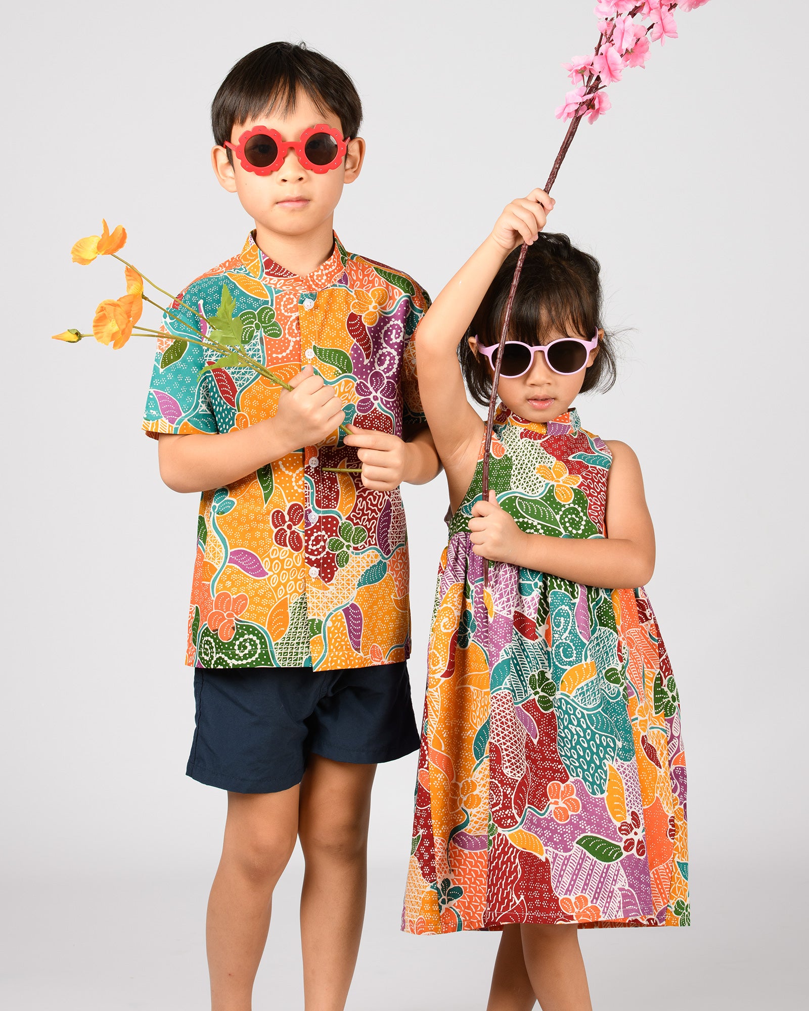 Two children in colorful, twinning batik outfits with floral patterns, holding flowers against a plain background.