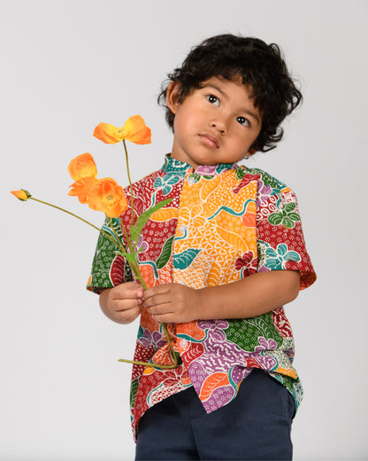 Child wearing a colorful batik patterned shirt holding flowers against a plain background