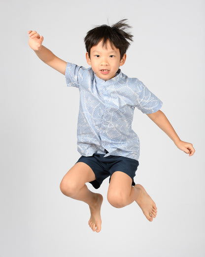 Child in a blue patterned batik shirt and dark shorts jumping against a light gray background