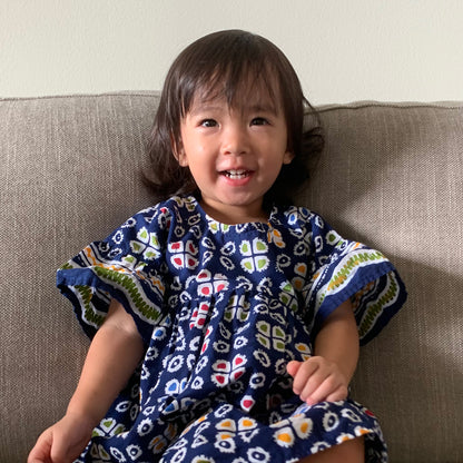 Smiling toddler sitting on a sofa wearing a navy blue batik dress with red yellow and green highlights