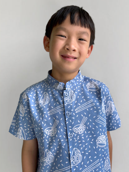 Boy wearing a blue batik shirt with bowl, chopsticks and rice pattern against a plain background